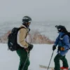Two skiers in winter gear standing on a snowy mountain slope, preparing for a run amidst a foggy landscape.