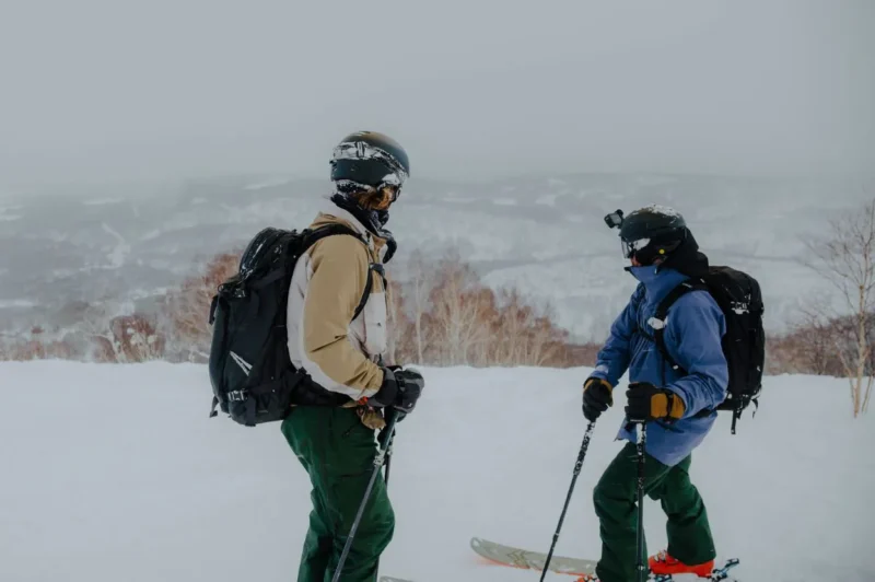 Two skiers in winter gear standing on a snowy mountain slope, preparing for a run amidst a foggy landscape.