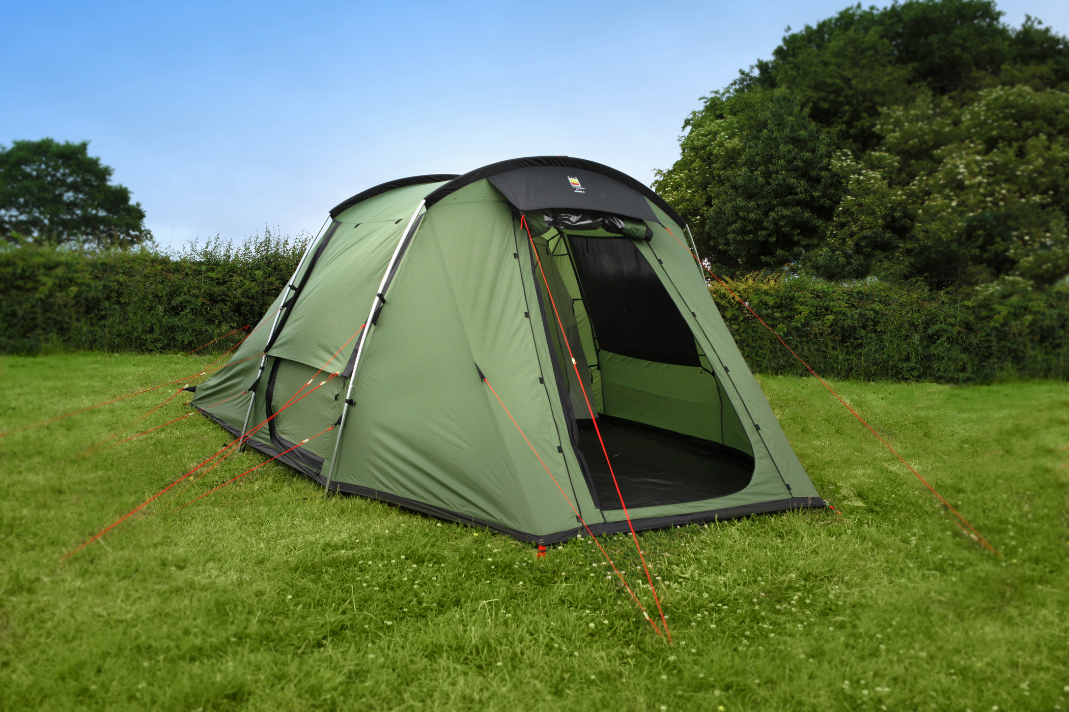 Green dome-style camping tent with its front door open, secured by bright orange guy lines and curved metal poles, pitched on a grassy field with a hedge and trees under a clear blue sky.