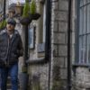 A man wearing a flat cap, checked shirt and dark puffer vest walks with his hands in his pockets down a narrow lane of old stone buildings adorned with hanging baskets and large mullioned windows.