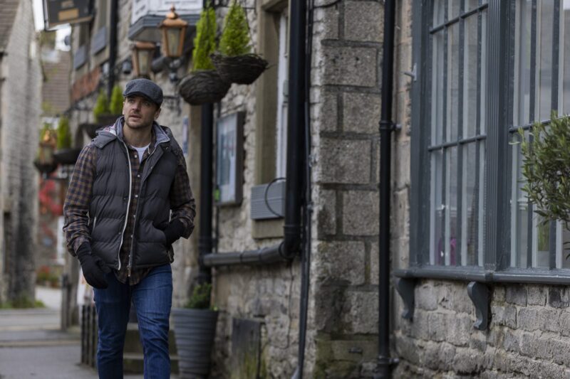 A man wearing a flat cap, checked shirt and dark puffer vest walks with his hands in his pockets down a narrow lane of old stone buildings adorned with hanging baskets and large mullioned windows.