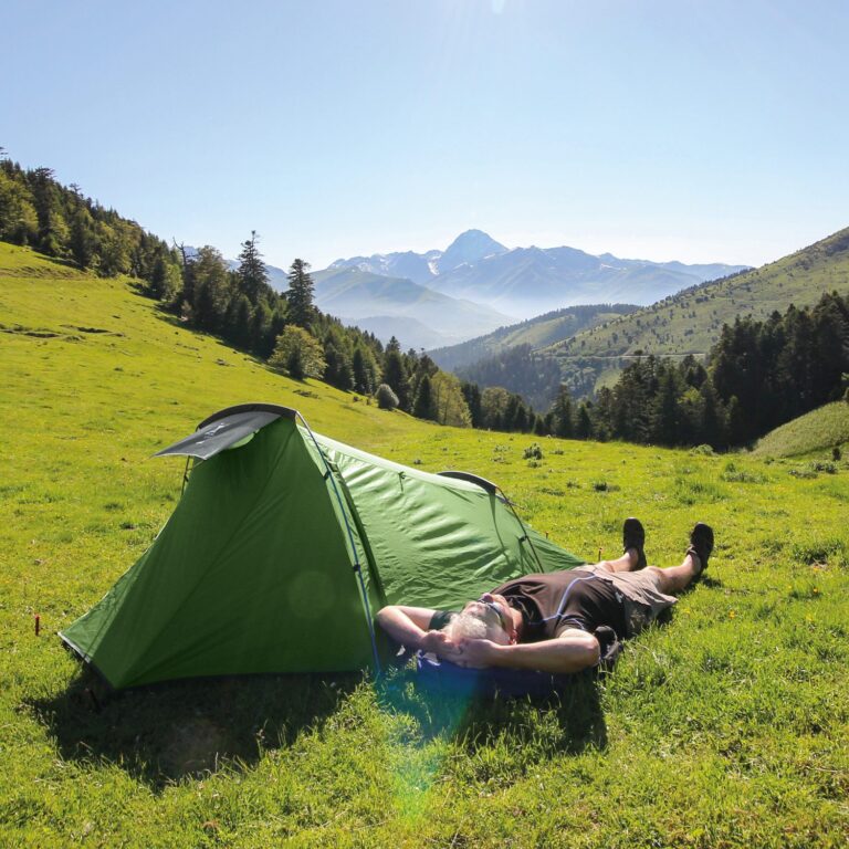 Person lying on their back with hands behind their head beside a bright green tent on a sunlit grassy slope, with tree-lined hills and distant snow-capped mountains under a clear blue sky