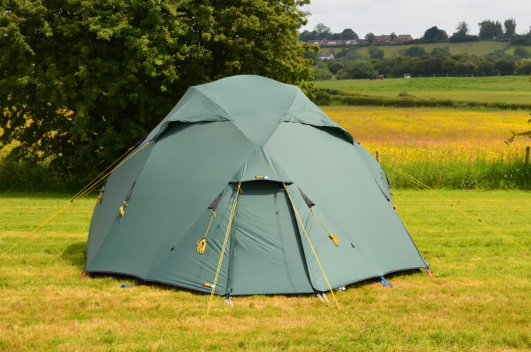 Dome-shaped green camping tent with yellow guy lines staked on a grassy field, backed by a large tree and a distant rolling meadow with houses on the horizon.