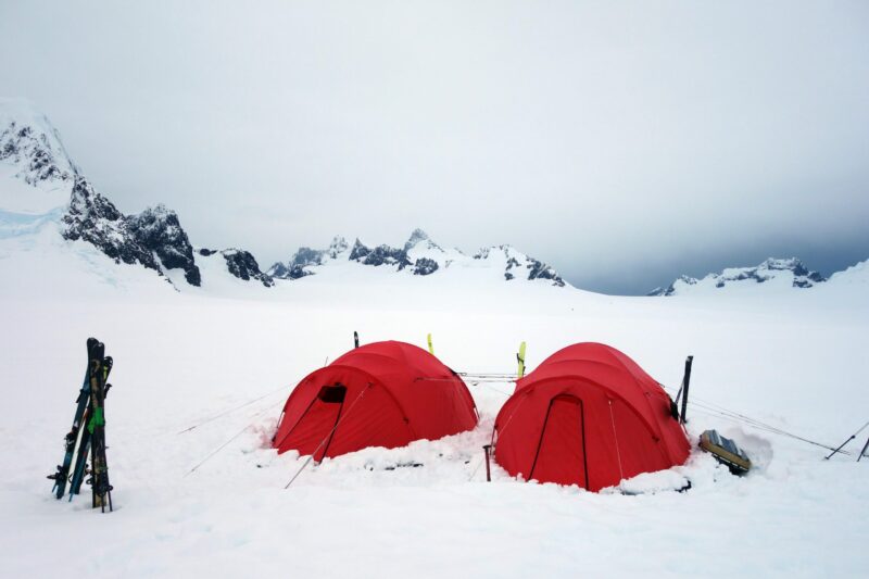 Two bright red dome tents pitched in deep snow on a windswept polar plain, with skis propped nearby and jagged, snow‑covered mountains under a grey overcast sky in the background.