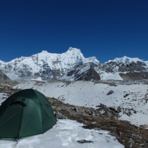 Green dome tent pitched on a snowy rocky high-altitude plain with expansive snow-covered jagged mountain peaks and glaciers beneath a clear deep-blue sky.