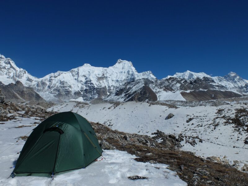 Green dome tent pitched on a snowy rocky high-altitude plain with expansive snow-covered jagged mountain peaks and glaciers beneath a clear deep-blue sky.
