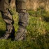 Lower legs and leather hiking boots covered by olive-green gaiters and outdoor trousers standing on a sunlit grassy meadow.