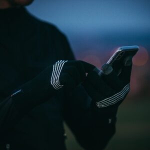 Close-up of hands wearing black knit touchscreen gloves with white stripe details tapping a smartphone, with a blurred dusk sky and bokeh city lights in the background.