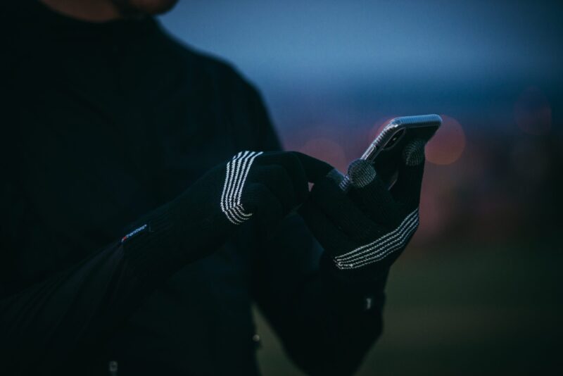 Close-up of hands wearing black knit touchscreen gloves with white stripe details tapping a smartphone, with a blurred dusk sky and bokeh city lights in the background.