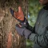 Person in a dark green jacket pulling on insulated black gloves with brown leather palms while standing beside a tree trunk in a blurred woodland background.