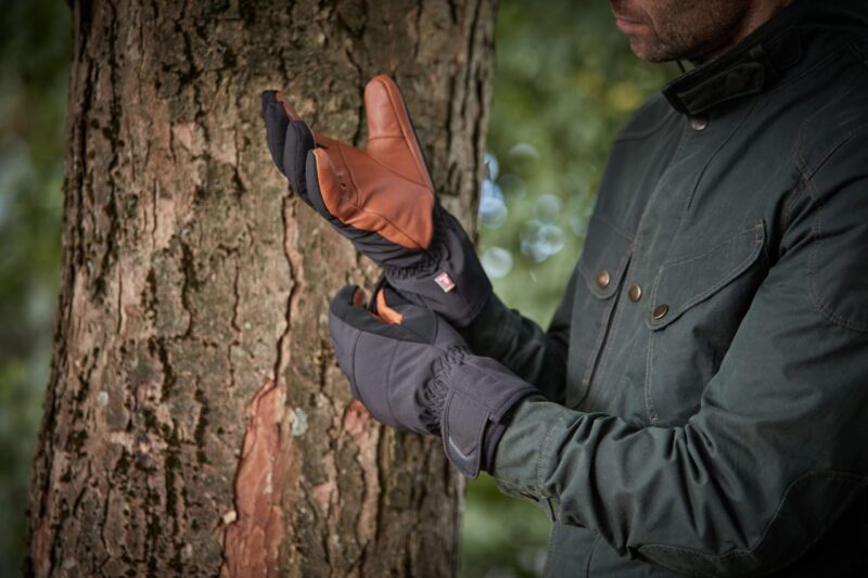 Person in a dark green jacket pulling on insulated black gloves with brown leather palms while standing beside a tree trunk in a blurred woodland background.