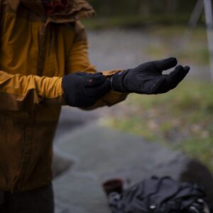 A person in a mustard-coloured jacket pulls on black insulated gloves outdoors, with a blurred grassy background and a backpack and mug on a nearby rock.