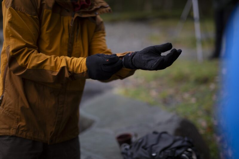 A person in a mustard-coloured jacket pulls on black insulated gloves outdoors, with a blurred grassy background and a backpack and mug on a nearby rock.