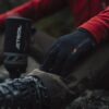 Close-up of a hiker's gloved hand wearing a black extremities glove resting on a muddy trouser knee, red jacket sleeve visible, with a hiking boot and blurred Jetboil stove nearby.