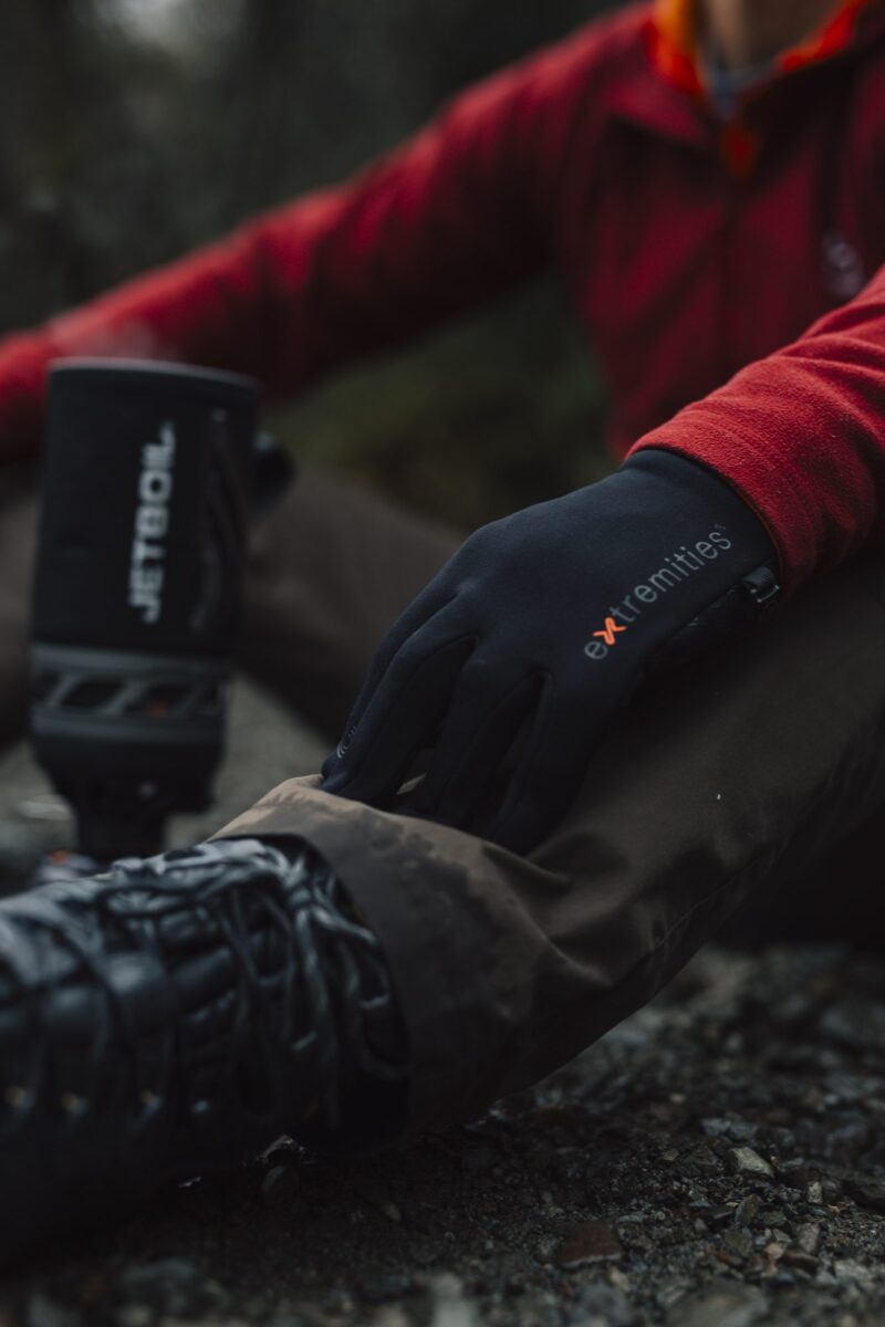 Close-up of a hiker's gloved hand wearing a black extremities glove resting on a muddy trouser knee, red jacket sleeve visible, with a hiking boot and blurred Jetboil stove nearby.