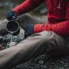 Person in a red fleece and black gloves sitting on a rocky riverbank, pouring water from a camping pot into a metal cup beside a small portable gas stove.