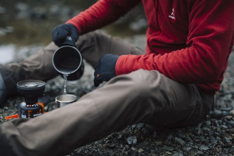 Person in a red fleece and black gloves sitting on a rocky riverbank, pouring water from a camping pot into a metal cup beside a small portable gas stove.