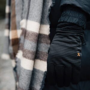 Close-up of a person outdoors near water wearing a black insulated glove with a small orange logo and a dark puffer sleeve, beside a grey, brown and cream striped wool scarf.