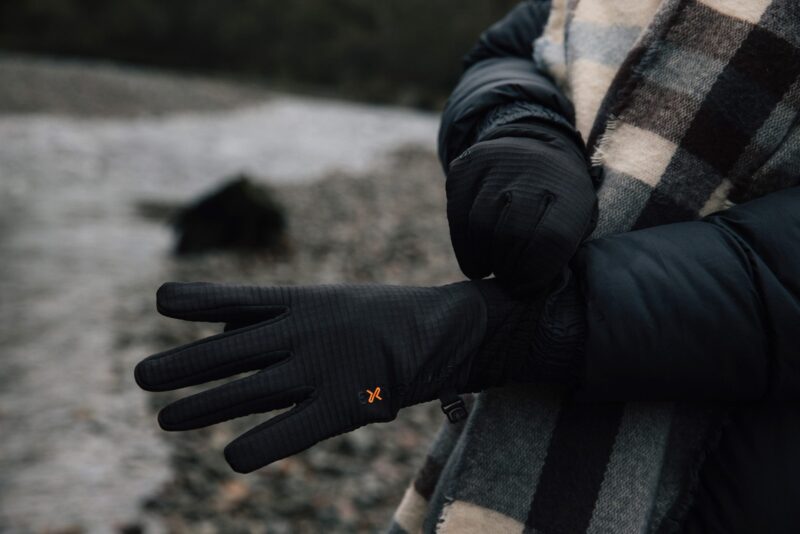 Person on a stony shoreline wearing a dark puffer jacket and checked scarf adjusts a pair of black insulated gloves with a small orange logo.