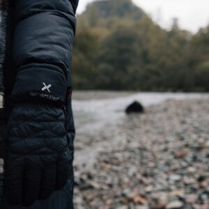 Person's arm in a black puffer coat wearing a black quilted Extremities glove next to a grey-and-cream striped scarf, standing on a pebble riverbank with trees blurred in the background.
