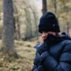A person in a black beanie, dark padded winter jacket and gloves pulls the collar up around their face and looks down while standing in a mossy, tree-filled forest.
