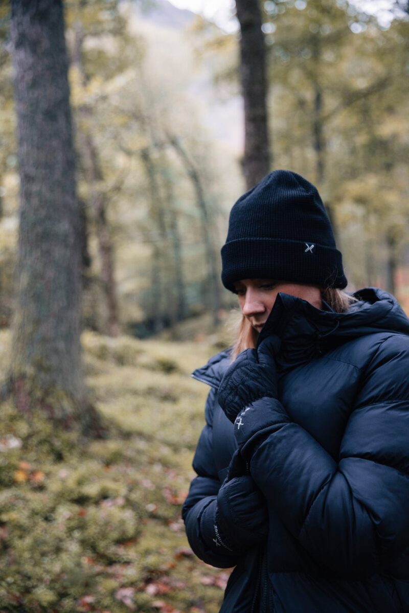 A person in a black beanie, dark padded winter jacket and gloves pulls the collar up around their face and looks down while standing in a mossy, tree-filled forest.