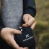 Close-up of a person’s hands holding a small black drawstring pouch stamped with a white extremities logo, the person wearing a grey sweater and dark jacket against a blurred grassy background.