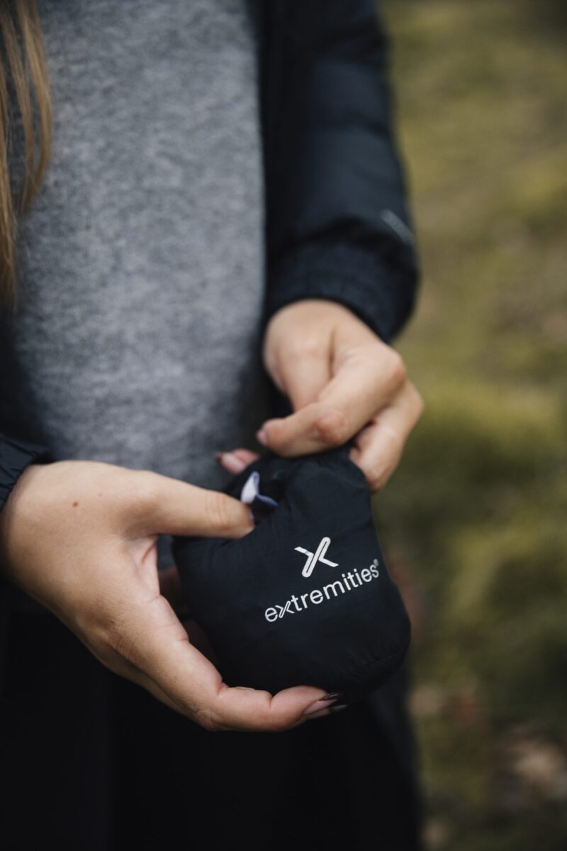 Close-up of a person’s hands holding a small black drawstring pouch stamped with a white extremities logo, the person wearing a grey sweater and dark jacket against a blurred grassy background.