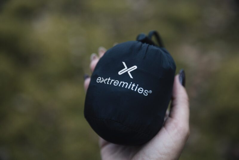 Hand holding a small black drawstring stuff sack stamped with a white "extremities" logo against a blurred green outdoor background.