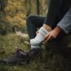 Person sitting on a mossy log in an autumn woodland, pulling a thick grey hiking sock onto one foot while maroon hiking boots rest on the grass beside them.