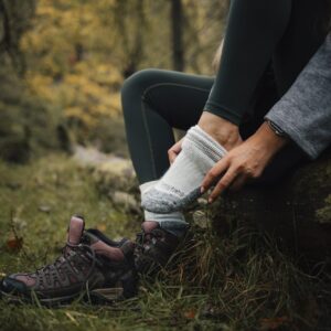 Person sitting on a mossy log in an autumn woodland, pulling a thick grey hiking sock onto one foot while maroon hiking boots rest on the grass beside them.