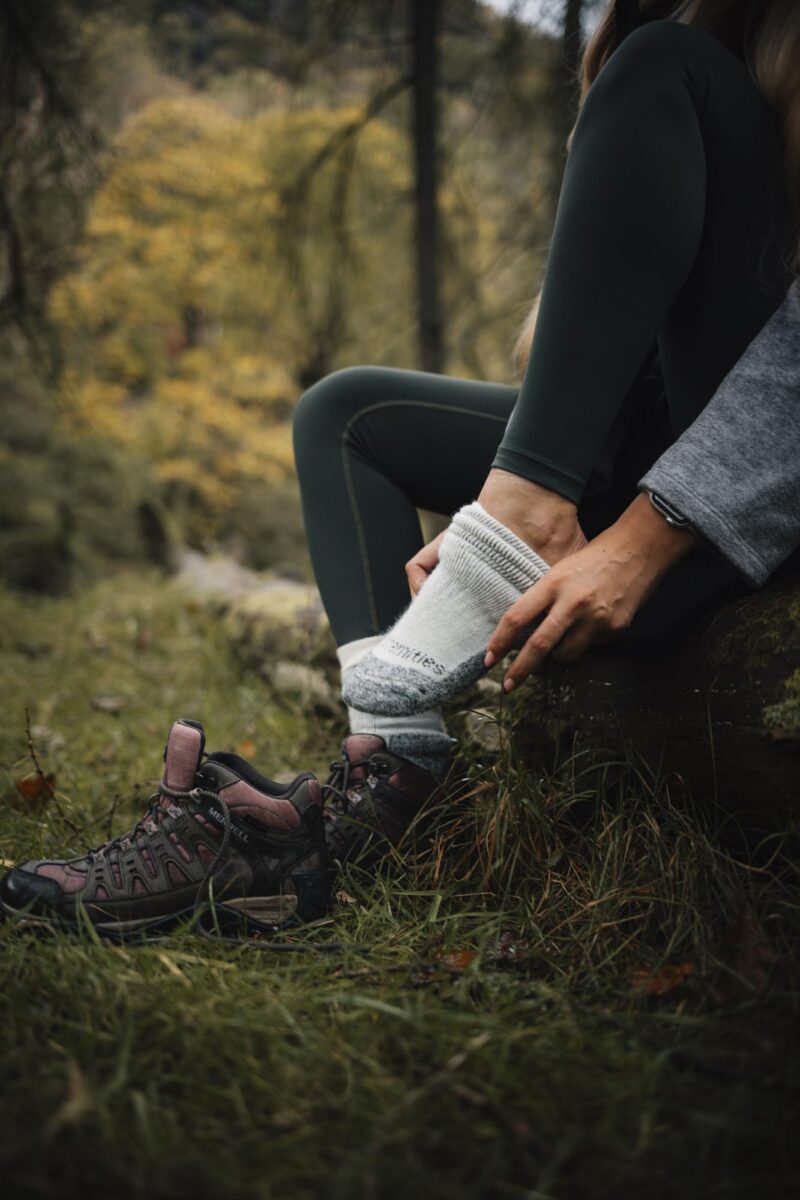 Person sitting on a mossy log in an autumn woodland, pulling a thick grey hiking sock onto one foot while maroon hiking boots rest on the grass beside them.