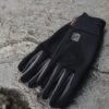 Black lightweight gloves with grey fingertip panels and a small logo, laid flat on a rough grey stone surface