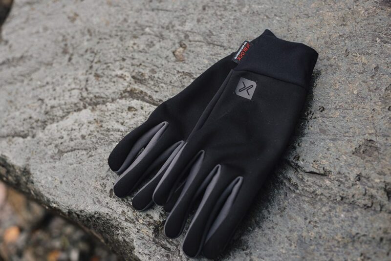 Black lightweight gloves with grey fingertip panels and a small logo, laid flat on a rough grey stone surface