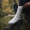 Close-up of a person pulling a thick white wool hiking sock over dark green leggings with their foot resting on a brown hiking shoe in a grassy, autumnal forest setting.