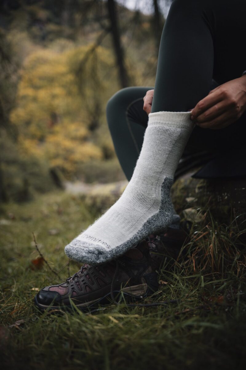 Close-up of a person pulling a thick white wool hiking sock over dark green leggings with their foot resting on a brown hiking shoe in a grassy, autumnal forest setting.