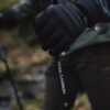 Close-up of a black-gloved hand gripping the handle of a carbon trekking pole labelled TRAIL CARBON, with a blurred woodland background and a hiker in outdoor clothing.