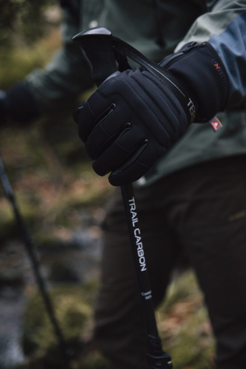 Close-up of a black-gloved hand gripping the handle of a carbon trekking pole labelled TRAIL CARBON, with a blurred woodland background and a hiker in outdoor clothing.