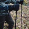 Close-up of a hiker's hands in black insulated gloves gripping Terra Nova carbon trekking poles, wearing a green jacket and brown trousers while standing on a mossy, leaf-strewn forest floor.