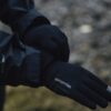 Close-up of a person’s crossed hands wearing black Extremities touchscreen gloves with a grey fingertip and a black Hydroshell jacket sleeve against a blurred outdoor ground.