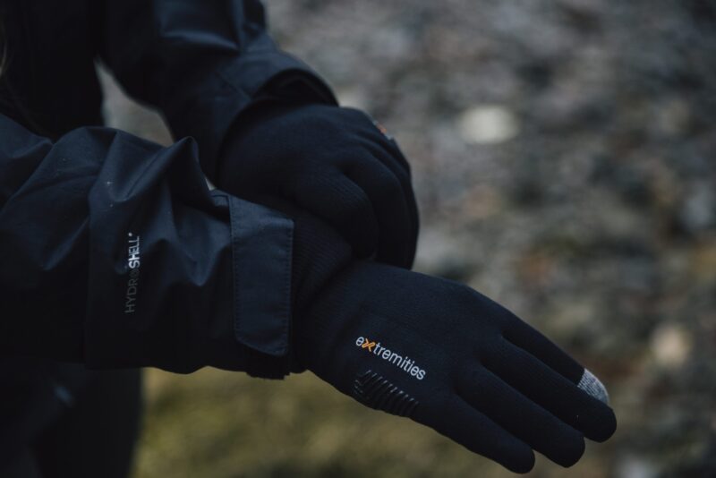 Close-up of a person’s crossed hands wearing black Extremities touchscreen gloves with a grey fingertip and a black Hydroshell jacket sleeve against a blurred outdoor ground.