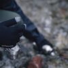 Close-up of a gloved hand wearing black Extremities touchscreen gloves with grey fingertips holding a small stainless-steel camping cup above a rocky, out-of-focus ground with a metal water bottle blurred in the background.