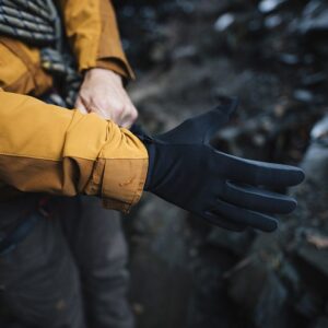 Person outdoors pulling a black glove onto their hand over a mustard-yellow jacket sleeve, rocky ground visible in the background.