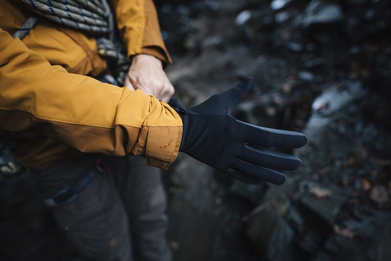 Person outdoors pulling a black glove onto their hand over a mustard-yellow jacket sleeve, rocky ground visible in the background.