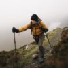 Hiker in a mustard-coloured jacket and black beanie climbs a foggy mountain trail with trekking poles and a backpack, a pale lake visible through the mist below.
