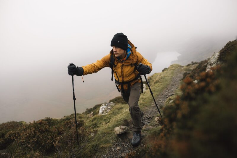 Hiker in a mustard-coloured jacket and black beanie climbs a foggy mountain trail with trekking poles and a backpack, a pale lake visible through the mist below.
