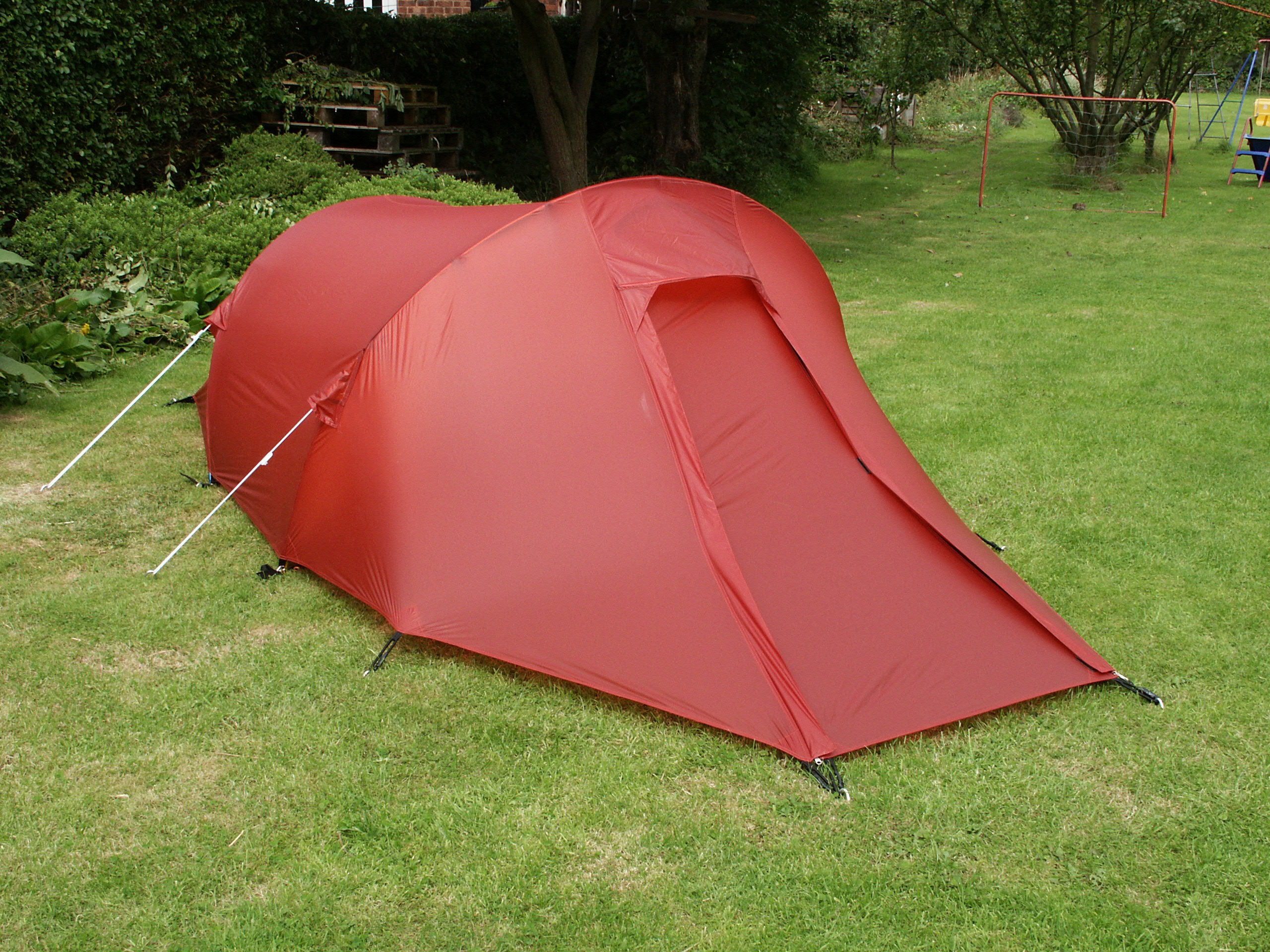 Red tunnel-style tent pitched on a mown lawn in a backyard, secured with white guylines and stakes, with shrubs, trees and children's play equipment visible in the background.