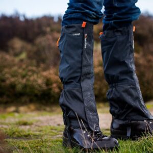 Close-up of lower legs wearing black waterproof gaiters over hiking boots and blue jeans, water droplets beading on the fabric as the wearer stands on grassy, muddy ground with blurred heathland behind,
