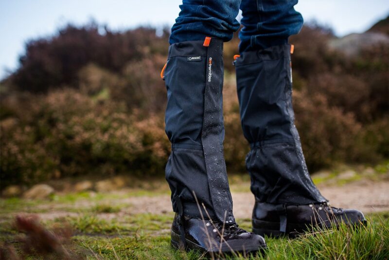 Close-up of lower legs wearing black waterproof gaiters over hiking boots and blue jeans, water droplets beading on the fabric as the wearer stands on grassy, muddy ground with blurred heathland behind,