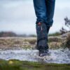 Lower legs in blue jeans and black waterproof gaiters and boots splashing through a muddy puddle on a windswept moorland path, water droplets frozen mid‑air against an overcast sky.
